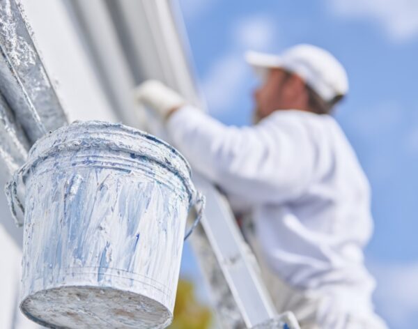A person in white is painting the exterior of a house using a ladder and a bucket on the roof, in a close-up view. --ar 1:1 --profile hkenas3 --v 7 Job ID: 6293bf11-b692-48dc-bdb0-199437ff9c50